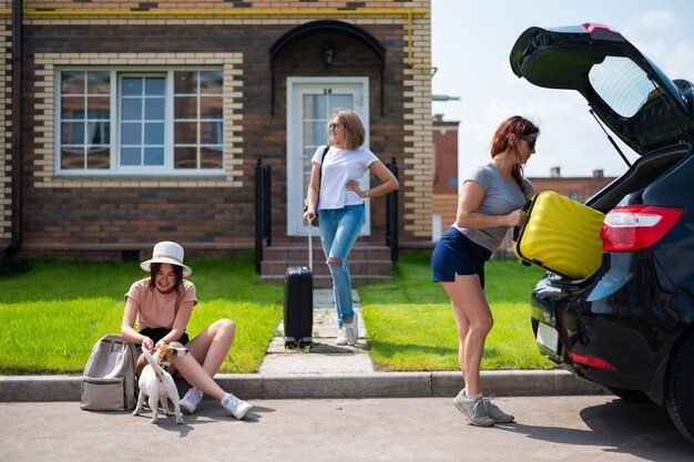 Three women outside a house load a car and luggage; one pets a small dog on the curb, another stands by suitcases.