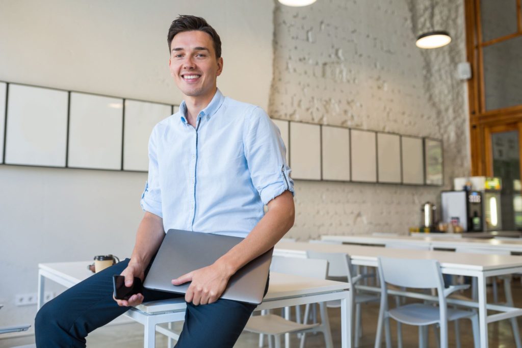 Smiling man in a light blue shirt sits on a table in a modern office, holding a laptop and phone.