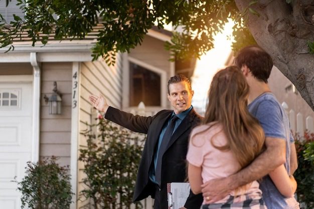 Real estate agent gestures toward a house while speaking to a couple standing arm in arm beneath a tree at sunset.