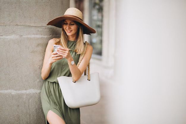 Woman in green dress and hat using phone, carrying a white tote bag by a textured wall.


